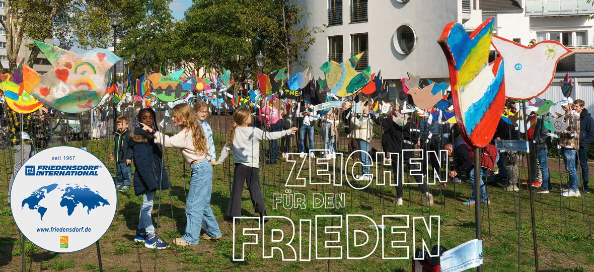 Das Bild zeigt eine Vielzahl Kinder mit bunt bemalten Friedenstauben an langen Stecken, welche auf einem Rasen vor einem Gebäude eingesteckt sind. Dazu ist ein Sticker des Friedensdorfes zu sehen und der Text: „Zeichen für den Frieden“