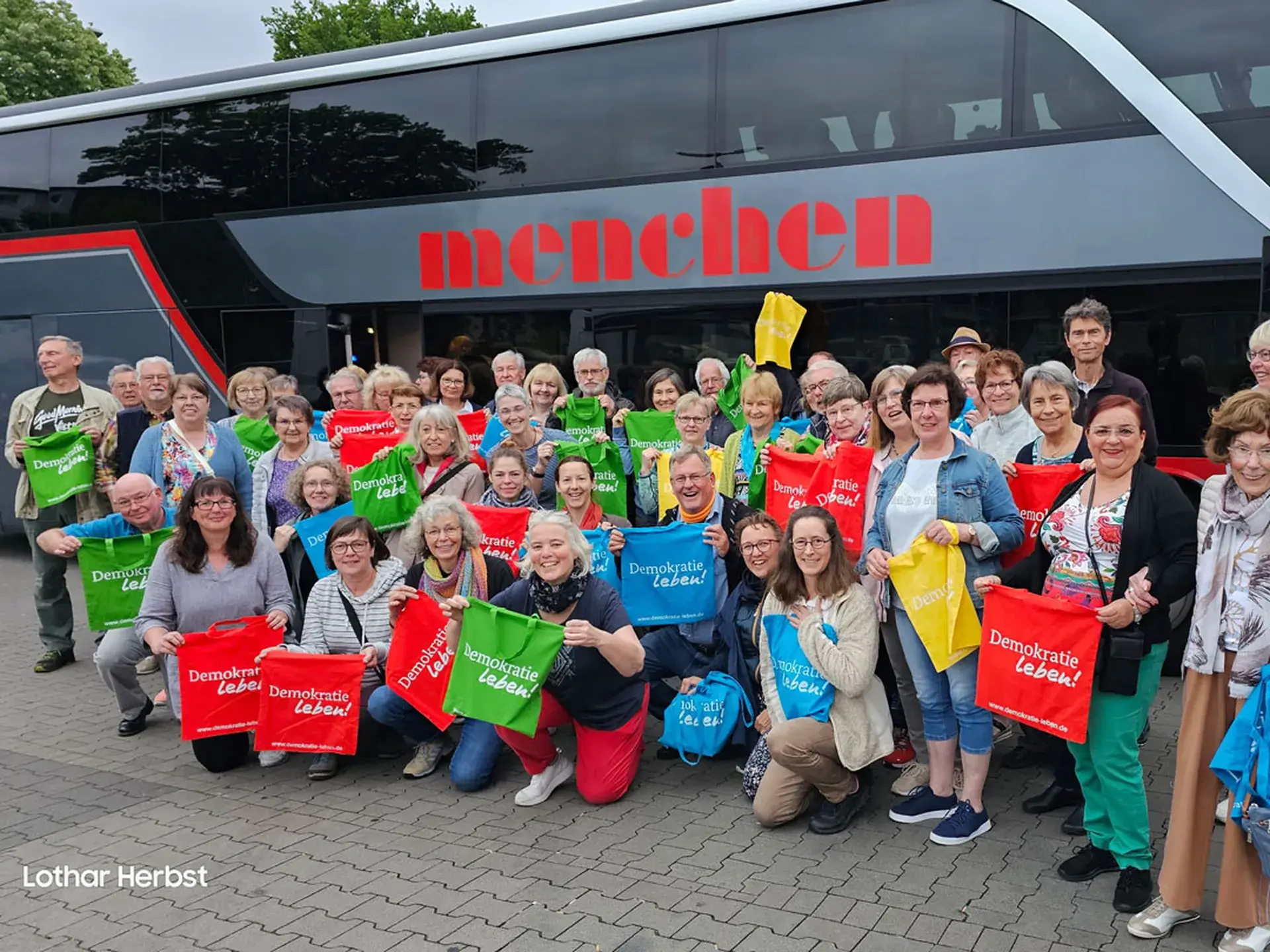 Reisebus / Gruppenbild der Reisegruppe vor ihrer Abreise. Alle halten bunte Stoffbeutel mit dem „Demokratie Leben“ Schriftzug in den Händen.